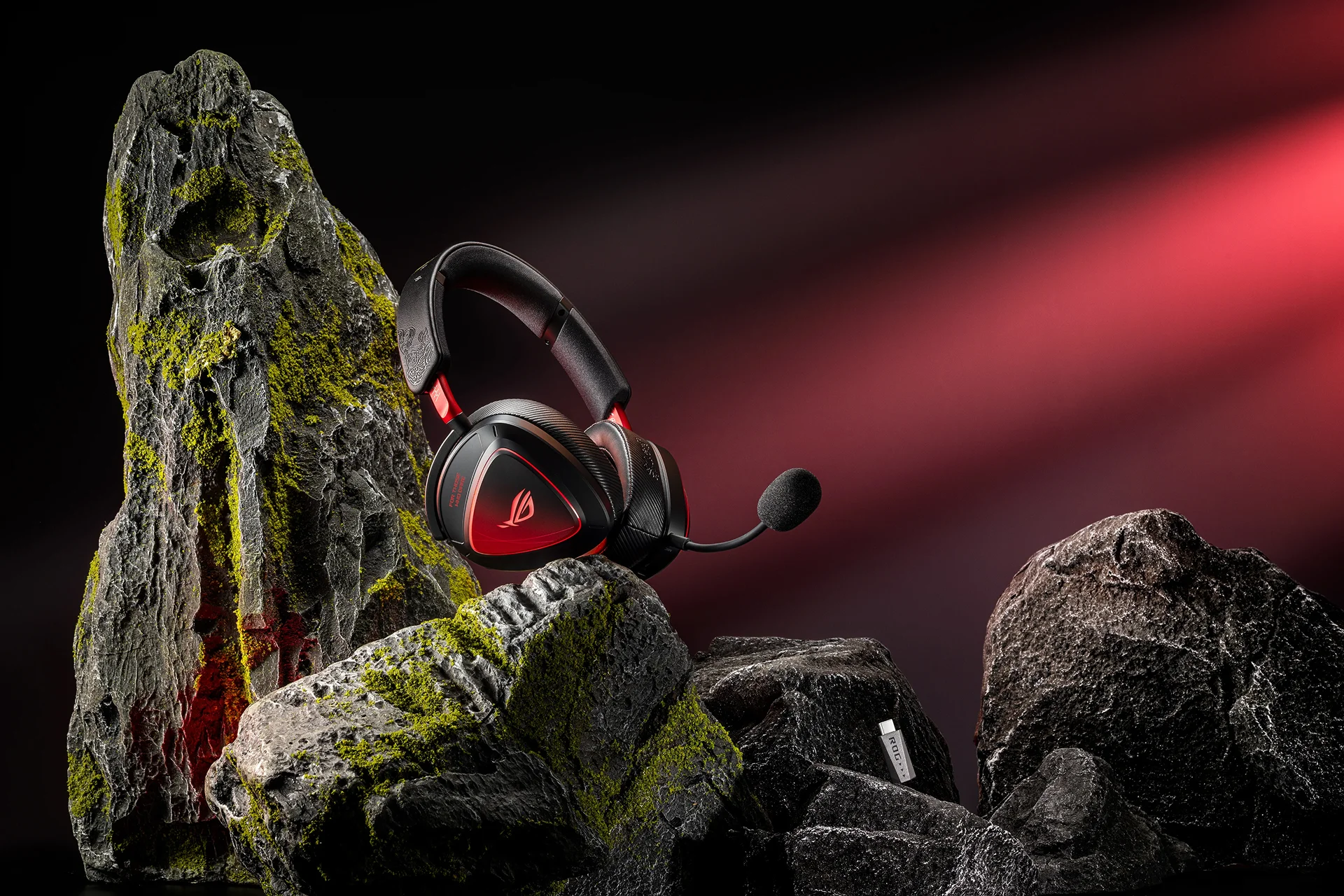 Close-up of ROG x Phantom Blade Zero gaming headset with a microphone and a dongle, displayed on mossy rocks with dramatic red lighting