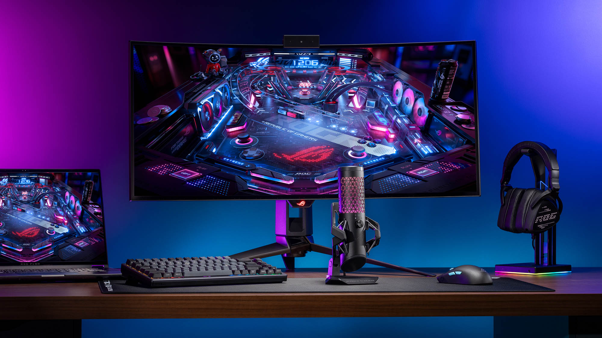 The ROG Carnyx microphone sitting on a desk in front of a gaming monitor, laptop, and headset.
