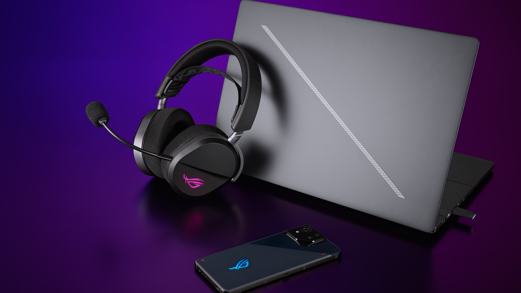 ROG Pelta gaming headset leaning against a ROG Zephyrus laptop beside a ROG Phone 9.
