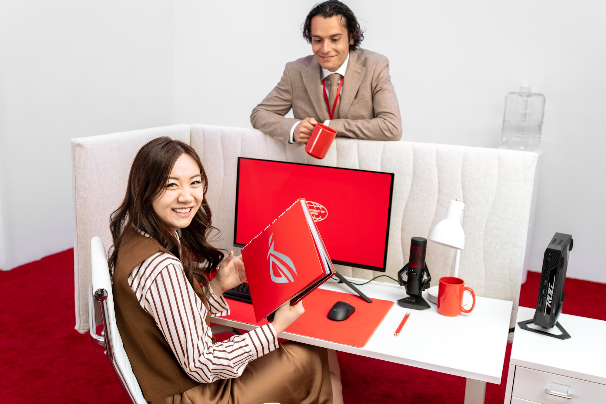 A woman sits at an office desk holding an ROG book while a young man with a coffee cup leans on the dividing wall of the cubicle