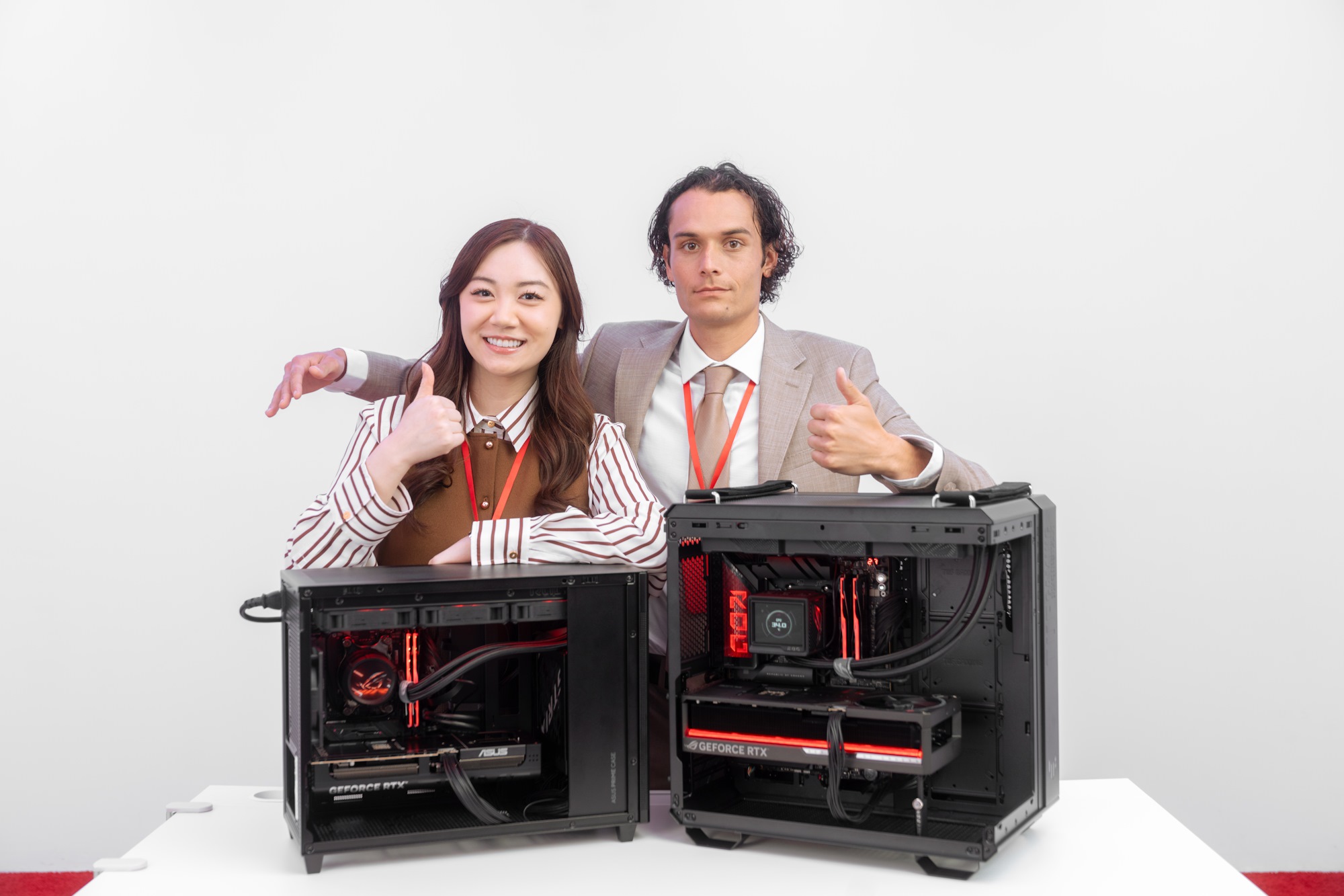 A man and a woman stand behind two completed PC builds, both giving a thumbs up, with the man extending his arm around the woman's shoulder without actually touching her