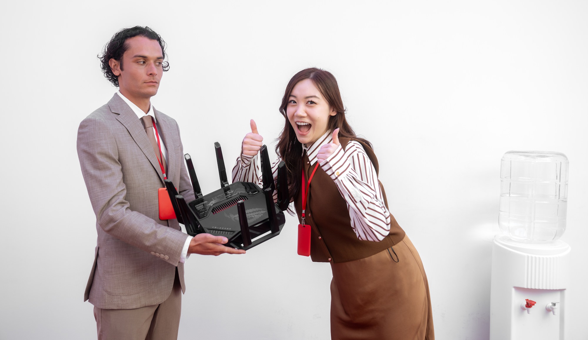 A man and a woman stand by a water cooler, the man holding a wireless router while the woman gives an enthusiastic two thumbs up