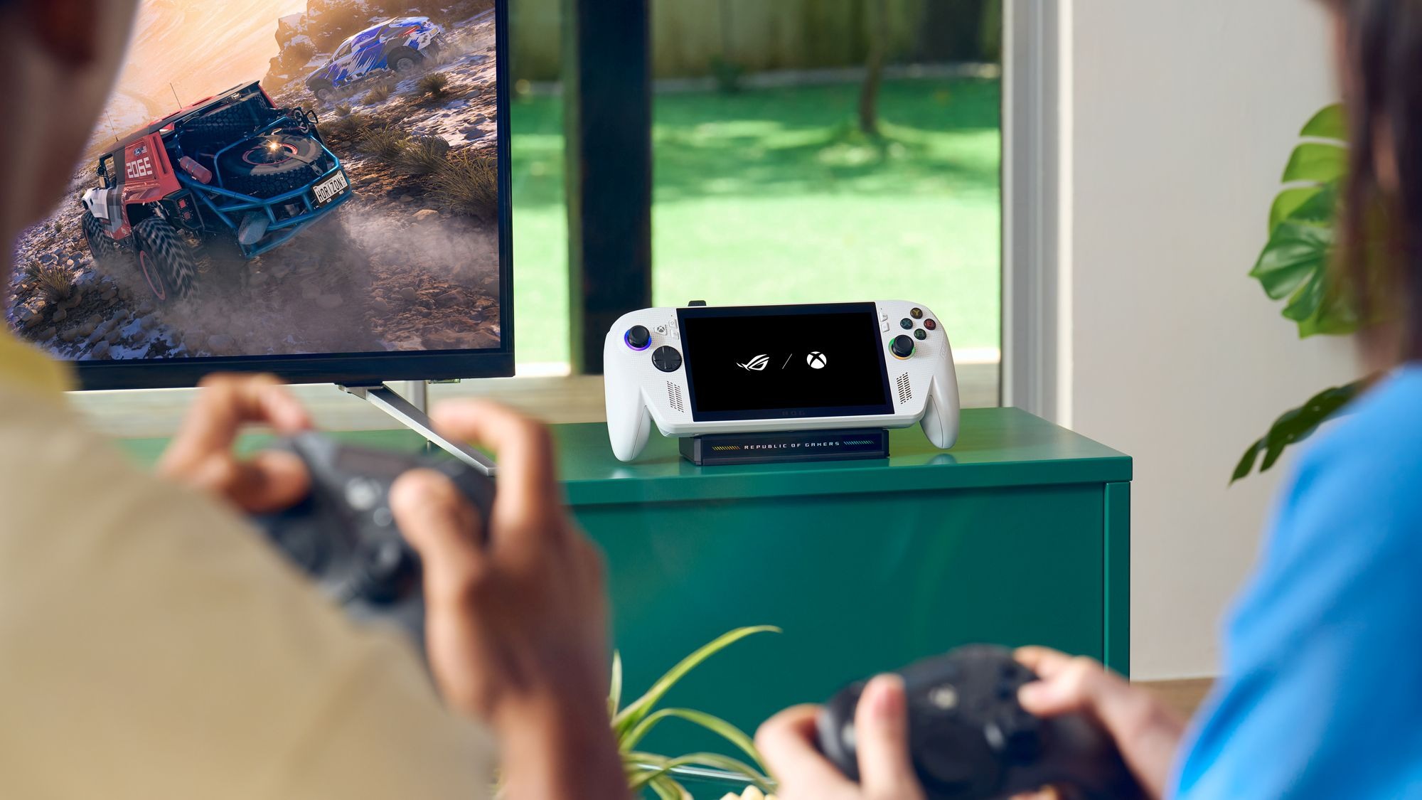 The ROG Xbox Ally sitting on a dock next to a TV, with two people playing a game on-screen.