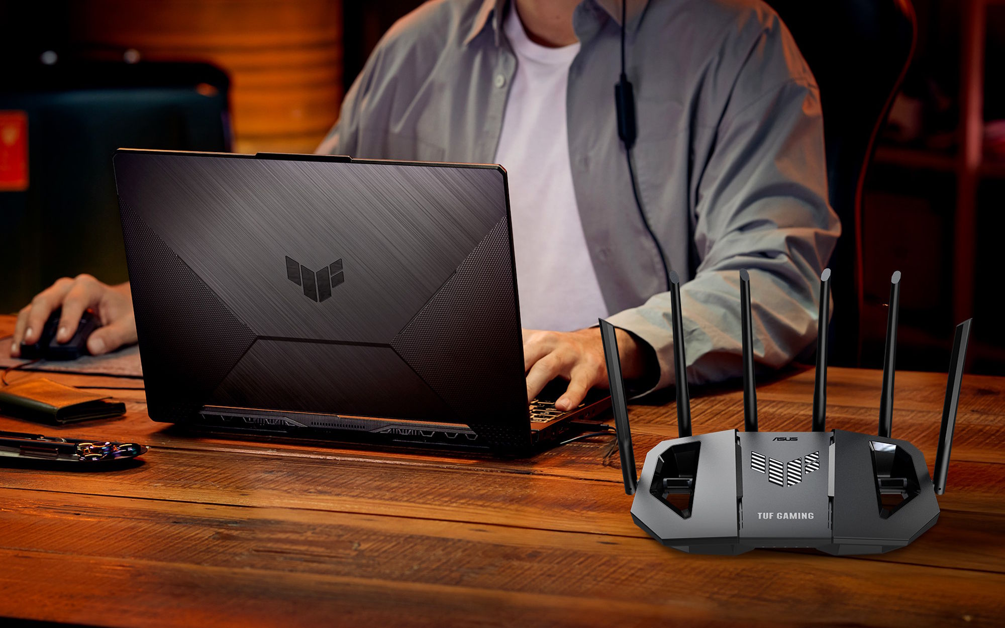 A gamer sits at a wooden desk playing PC games with a TUF Gaming laptop, peripherals, and the TUF Gaming BE6500 wireless router