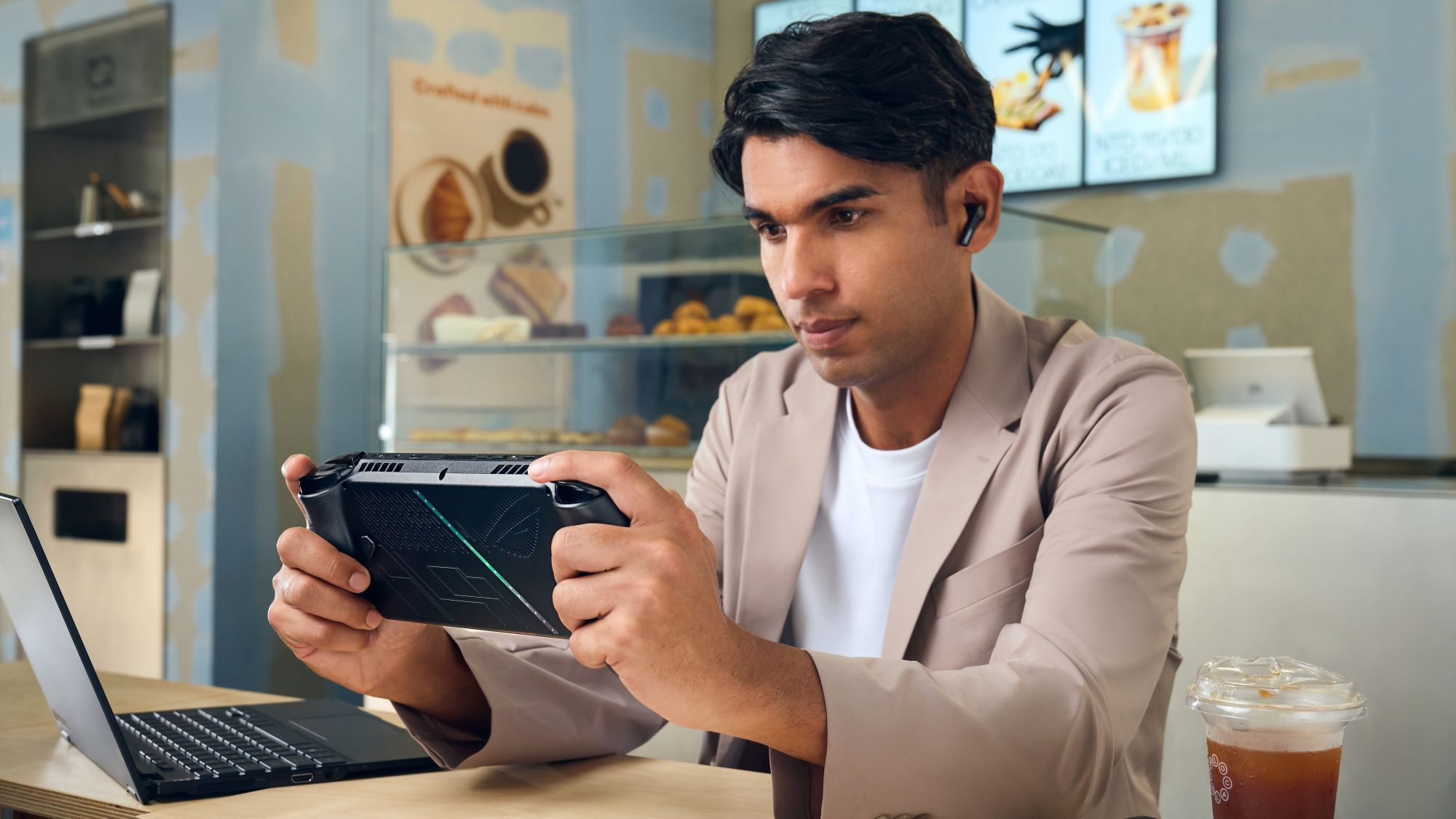 A man plays on an ROG Xbox Ally X while seated beside a laptop and beverage.