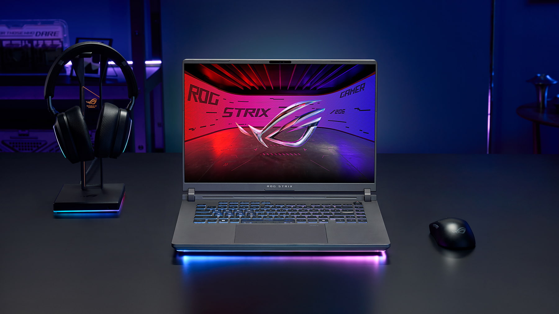 Notebook ROG Strix G16 aberto sobre uma mesa escura, acompanhado de um headset em suporte iluminado e um mouse gamer, em um setup com luzes azuis e roxas.