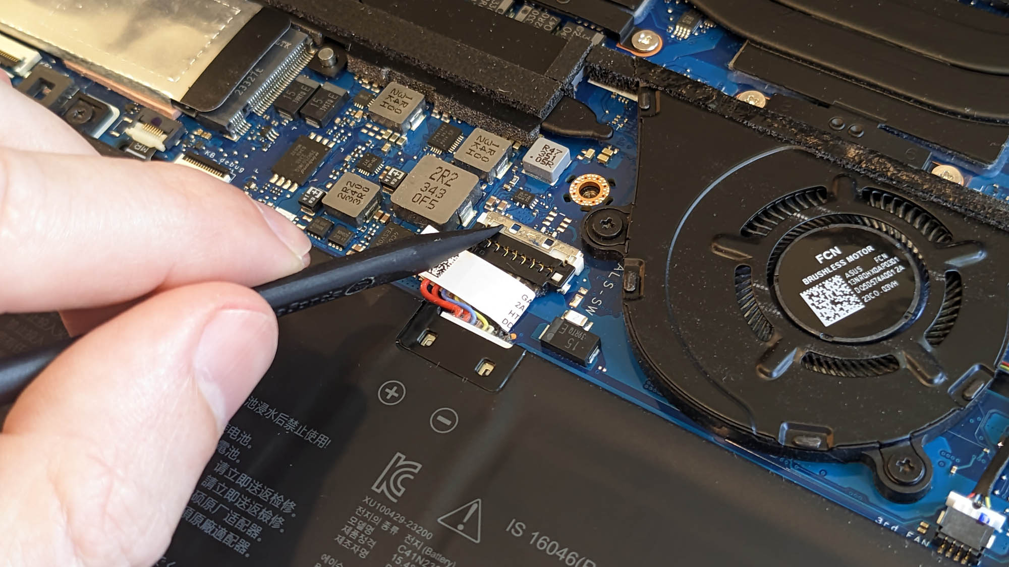 A hand pushing a plastic tool against the battery retaining clip of a Zephyrus G14 laptop